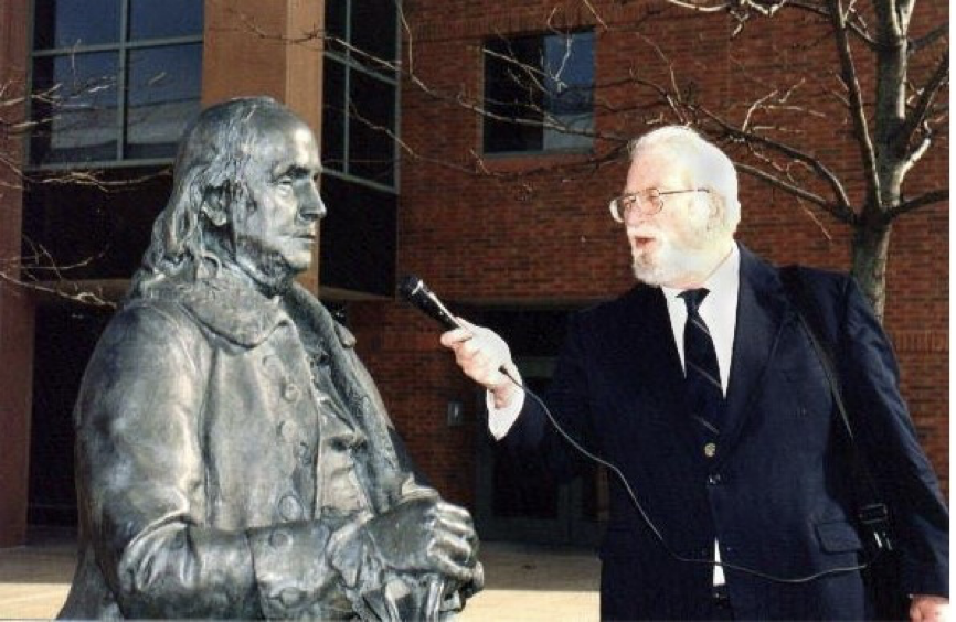 Charles Stuart Kennedy holds up microphone to interview a statue of Ben Franklin, often considered our first diplomat