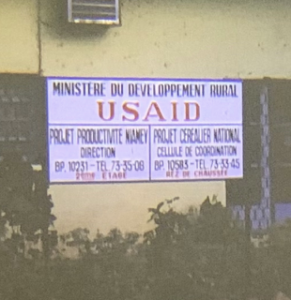 Sign posted on side of building. "Minestere du Development Rural USAID" Project Productive Niamey. Contact info.